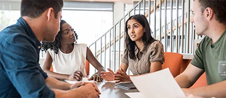 Four people sit around a table in a bright, modern workspace engaged in discussion. One person speaks while the others listen attentively. Papers and an open document are visible on the table.