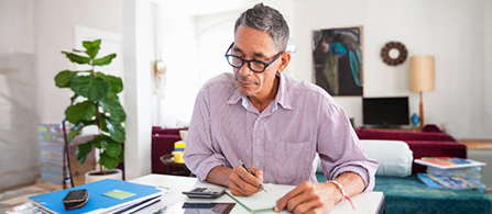A person with gray hair and glasses, wearing a checked shirt, sits at a table writing in a notebook in a living room with a sofa, plant, and lamp in the background.