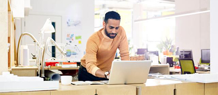 A person with a beard wearing an orange sweater stands leaning over a desk working on a laptop in a bright open office with desk lamps and supplies around them.