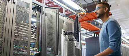 A person in a gray sweater and safety glasses stands operating a console beside rows of tall server cabinets with colored cabling, beneath orange overhead cable trays in a data center.