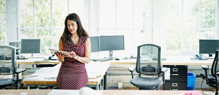 A person in a patterned dress stands in an open office reading a tablet, with empty desks, office chairs, and large windows around them.