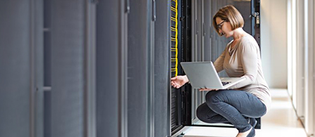 A person wearing glasses and a beige sweater crouches in a server room aisle, holding an open laptop while inspecting a server rack with yellow-highlighted equipment.