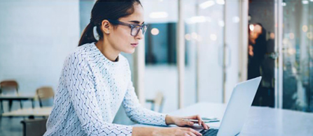 A person wearing glasses and a white patterned sweater sits at a desk in a modern open office, focused on typing on a laptop. Another person is blurred in the background.