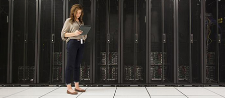 A person wearing a beige top and dark trousers stands in a long corridor between tall rows of black server racks in a data center, looking down at a tablet held in both hands.