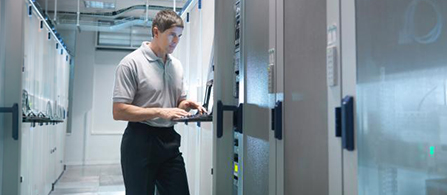 A person wearing a gray polo shirt stands in a brightly lit data center corridor, holding an open laptop and looking at a rack of server equipment to their side.