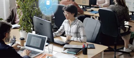 A person sits at a workstation in a busy open-plan office, smiling at a colleague across the desk. A digital security shield icon with a lock floats above the desk, suggesting a cybersecurity theme. A coffee cup is visible on the desk.