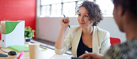 A person with curly hair wearing a white blazer sits at a desk holding a pen, looking attentively toward someone partially visible in the foreground. Office supplies, a small plant, and a red accent wall are visible in the background.