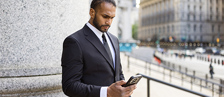 A person wearing a dark suit and tie stands outdoors leaning against a stone column, looking down at a smartphone with a focused expression. A busy city street and buildings are visible in the background.