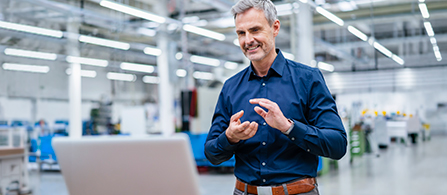 A person wearing a dark navy shirt stands smiling in a large, brightly lit industrial facility, gesturing while looking at an open laptop positioned on a surface in front of them. The warehouse space extends into the background.