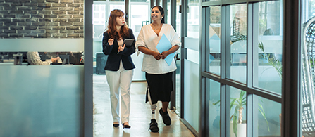 Two professionals walking and talking in a modern office hallway with large windows and natural light.
