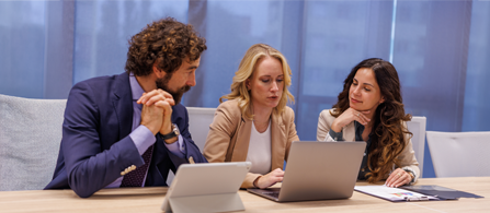 Three professionals sit together at a conference table, focusing on a laptop screen. The central woman types while the others listen and observe, suggesting a collaborative meeting or discussion. A tablet and documents lie on the table, and large blue curtains create a modern corporate backdrop. The overall scene conveys teamwork, concentration, and professional communication in an office environment.