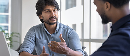Two people are seated facing each other in conversation in a bright modern office. One gestures expressively while speaking; the other listens attentively.
