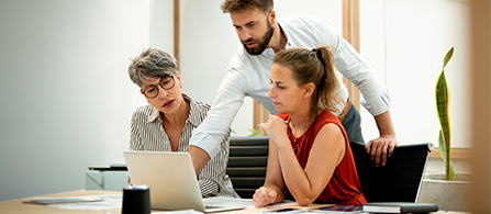 Three people collaborate around a laptop at a desk in an office. Two are seated and one stands leaning in, all focused on the laptop screen.
