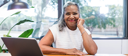 A person with gray hair sits at a wooden desk with an open laptop, resting their chin on one hand and smiling warmly at the camera. A plant and bright windows are visible in the background.