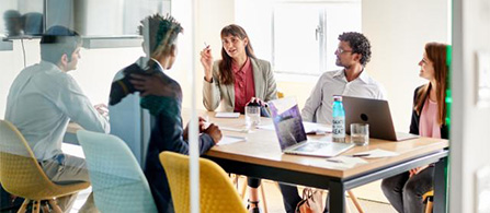 Five people sit around a conference table in a glass-walled meeting room in discussion. One person stands at the head of the table gesturing while others listen. Laptops, documents, and water bottles are on the table.