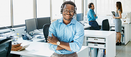 A person in a light blue button-down shirt and headset stands with arms crossed and a broad smile in a bright open-plan office. Two other people are visible conversing in the background.