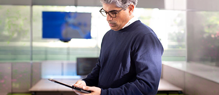 A person wearing glasses and a navy sweater stands in a modern control room, looking down at a tablet with a thoughtful expression. A large monitor displaying a map is visible in the background.