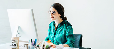 A person wearing glasses and a green polka-dot blouse sits at a desk in a bright office, looking intently at a large monitor. Pens and desk supplies are in the foreground, and the wall behind is plain white.