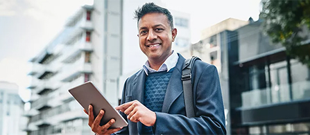 A person in a dark suit with a bag strap across the shoulder stands outdoors holding a tablet and smiling toward the camera. Modern multi-story buildings rise softly out of focus in the background.