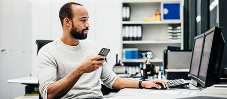 A person with a beard, wearing a light gray sweatshirt, sits at a desk holding a smartphone in one hand and resting the other on a keyboard while looking at a monitor. Shelves with binders line the bright office behind them.
