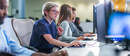 A person wearing glasses and a navy shirt sits focused at a computer in a busy operations center, one hand on the keyboard. A row of colleagues works at additional monitors along the same desk, receding into the background.