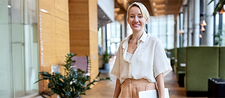 A person in a cream blouse and tan trousers stands smiling toward the camera in a bright, modern lobby with wood-paneled walls and potted plants. Soft daylight and blurred lounge seating fill the background.