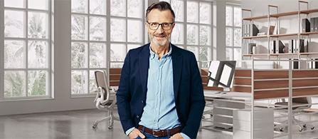 A person wearing glasses, a navy blazer over a light blue shirt, and jeans stands with hands in pockets, smiling in a bright office. Tall windows behind reveal greenery outside, with desks and shelving to the sides.