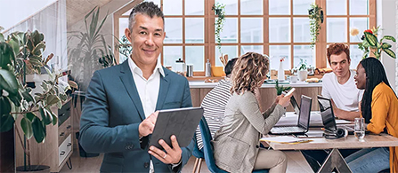 A person in a gray blazer holds a tablet and smiles toward the camera in the foreground of a plant-filled modern office. Behind them, three colleagues collaborate around a table with laptops and papers.
