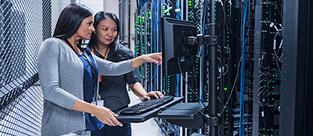 Two people stand together in a data center aisle. One in a gray sweater points at a monitor mounted on a rack while the other, in a dark top, works at a keyboard. Rows of server cabinets with cabling line both sides.