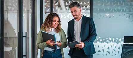 Two people walk and talk in a bright office corridor. One with curly hair, wearing a green cardigan, holds a folder; the other, in a dark blazer, holds papers. A patterned frosted-glass wall runs behind them.