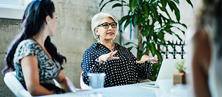 A person with short light hair and glasses, wearing a navy polka-dot top, gestures with both hands while speaking at a meeting table. Another person listens in the blurred foreground, and a leafy plant sits behind.