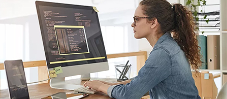 A person with hair in a ponytail, wearing glasses and a denim shirt, sits at a desk typing on a desktop computer whose screen displays lines of code on a dark background. Sticky notes frame the monitor and a bright, plant-filled office is visible behind.