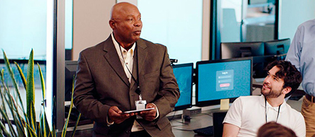 A person in a gray blazer holds a small device and talks with a colleague in a white shirt who is seated nearby in a security operations center. Monitors displaying dashboards glow in the background, and a plant sits in the foreground.