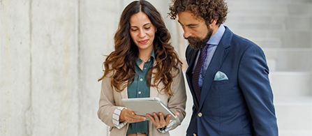 Two people stand outdoors beside a light stone wall, looking together at a tablet one of them holds. One wears a tan cardigan over a green blouse; the other wears a navy suit with a patterned tie and pocket square. Both look down at the screen.