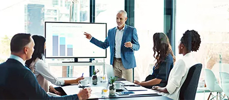 A person in a blue blazer stands and gestures toward a wall-mounted screen showing a bar chart while presenting to colleagues seated around a boardroom table. Tall windows with a city view brighten the room, and the seated people listen and take notes.