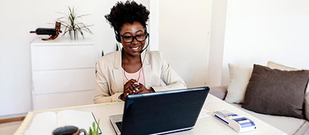 A person in a cream blazer wearing a headset sits at a desk in a bright home office, smiling at an open laptop during a video call with hands clasped. A couch, notebook, and small plant are visible in the background.