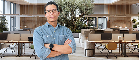 A person in a light blue button-down shirt and glasses stands with arms crossed and a slight smile in a bright, modern open-plan office. Rows of desks with monitors, plants, and wood-toned partitions are visible in the background.