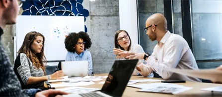 Four people sit around a conference table in a meeting, engaged in discussion. One person on the right gestures while speaking, and the others listen. Laptops, papers, and a whiteboard are visible in the bright office setting.