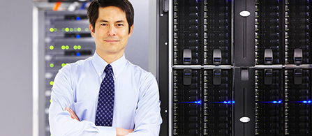 A person with short dark hair, wearing a light blue dress shirt and a patterned dark tie, stands with arms crossed in a data center and smiles toward the camera. Tall server racks with status lights fill the background on both sides.