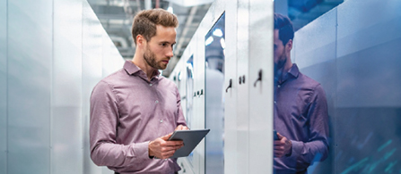 A person with short hair and a short beard, wearing a mauve button-up shirt, stands in a data center holding a tablet and looks toward the server cabinets with a focused expression. The equipment surfaces reflect blue light, and a reflection of the person is visible on a glossy panel.