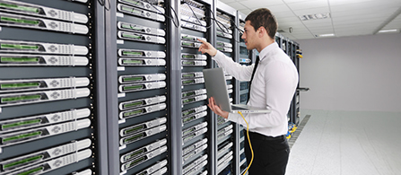 A person wearing a white dress shirt and dark tie stands in a data center holding an open laptop in one hand and reaching toward a tall server rack with the other, looking at the equipment. Rows of servers with green status lights line the aisle.