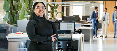 A person with dark hair pulled back, wearing glasses and a black turtleneck, stands with arms crossed in an open-plan office and smiles toward the camera. Behind them are desks with monitors and large potted plants, and three people in business attire walk together in the background.