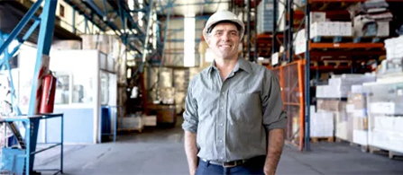 A person wearing a white hard hat and a gray button-up shirt stands in an industrial warehouse and smiles toward the camera. Behind them are metal shelving with stacked boxes, machinery, and a red fire extinguisher mounted on a column.