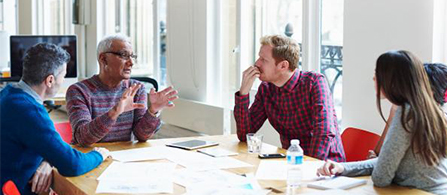 Four people sit around a table in a bright office in discussion. An older person in a patterned sweater gestures while speaking, and the others listen. Papers, a tablet, and water bottles are on the table.