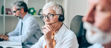 A person with short gray hair and glasses, wearing a headset and a white shirt, sits at a desk with a focused expression, resting a hand near their chin. Another person works at a desk in the background, and a third is partially visible in the blurred foreground.