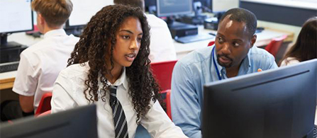 Two people sit side by side at computers in a classroom or training lab. One in a white shirt and tie looks at the monitor while the other, wearing a lanyard, leans in and points toward the screen. Other people work at computers in the background.