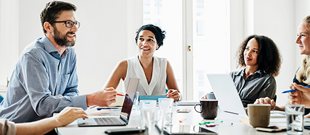 Four people sit around a table in a bright meeting room, smiling and engaged in a relaxed discussion. Laptops, notebooks, and coffee cups are on the table. One person on the left holds a pen and gestures while talking.