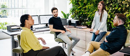 Four people gather in a casual seating area in a modern office surrounded by lush green plants, engaged in relaxed conversation. Some are seated on stools and chairs while others stand. Computers and office furniture are visible in the background.