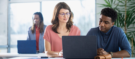 Two people sit side by side at a desk looking at a laptop screen together in a bright office. One wears a coral-colored top and glasses and points at the screen; the other wears a blue shirt and listens. A third person works in the background.