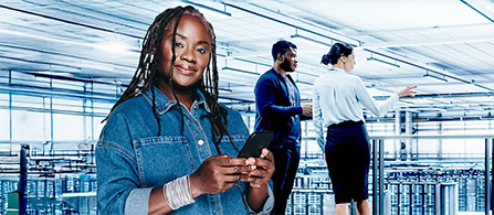 A person in a denim jacket stands in the foreground of a data center holding a smartphone and looking toward the camera. Two other people stand among the server racks in the background, one gesturing toward the equipment.
